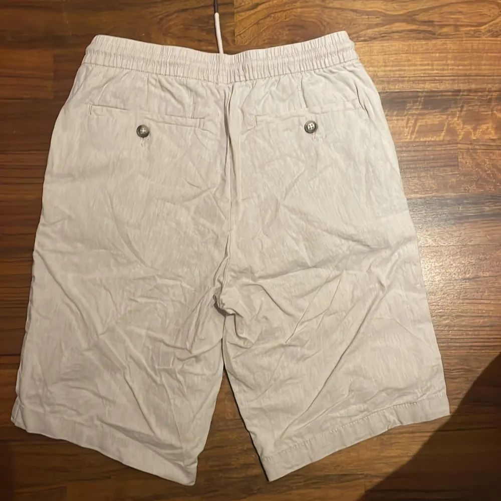 Säljer ett par bekväma beige shorts från Dressmann i storlek S. De är gjorda av en linneblandning och har en elastisk midja med dragsko för justerbar passform. Shortsen har två sidofickor och två bakfickor med knappar. Perfekta för varma sommardagar!. Shorts.