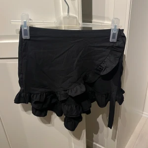 Svart kjol från BikBok - En svart kjol från BikBok med volangdetaljer. Kjolen är kort och har en omlott-design. Tillverkad av 100% polyester.