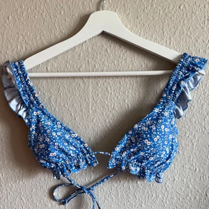 Blå blommig bikiniöverdel med volang - Säljer en superfin blå bikiniöverdel med små vita blommor. Den har volangdetaljer på axelbanden och knytning fram. Perfekt för sommarens alla badäventyr! 💙