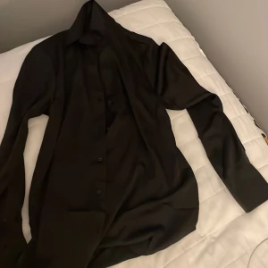 Svart skjorta från H&M - Säljer en svart skjorta från H&M i storlek S. Skjortan är tillverkad av 96% polyester och 4% elastan, vilket gör den både bekväm och stilig. den har en v- ringad krage och tyget har lite glans.  Den har långa ärmar och knappar framtill. Perfekt för både vardag och fest! 