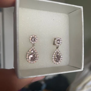 Glittriga örhängen med rosa stenar - Säljer ett par superfina glittriga örhängen med rosa stenar. De har en rund sten upptill och en droppformad sten nedtill, båda omgivna av mindre stenar som ger extra bling. Perfekta för fest eller bal! ✨