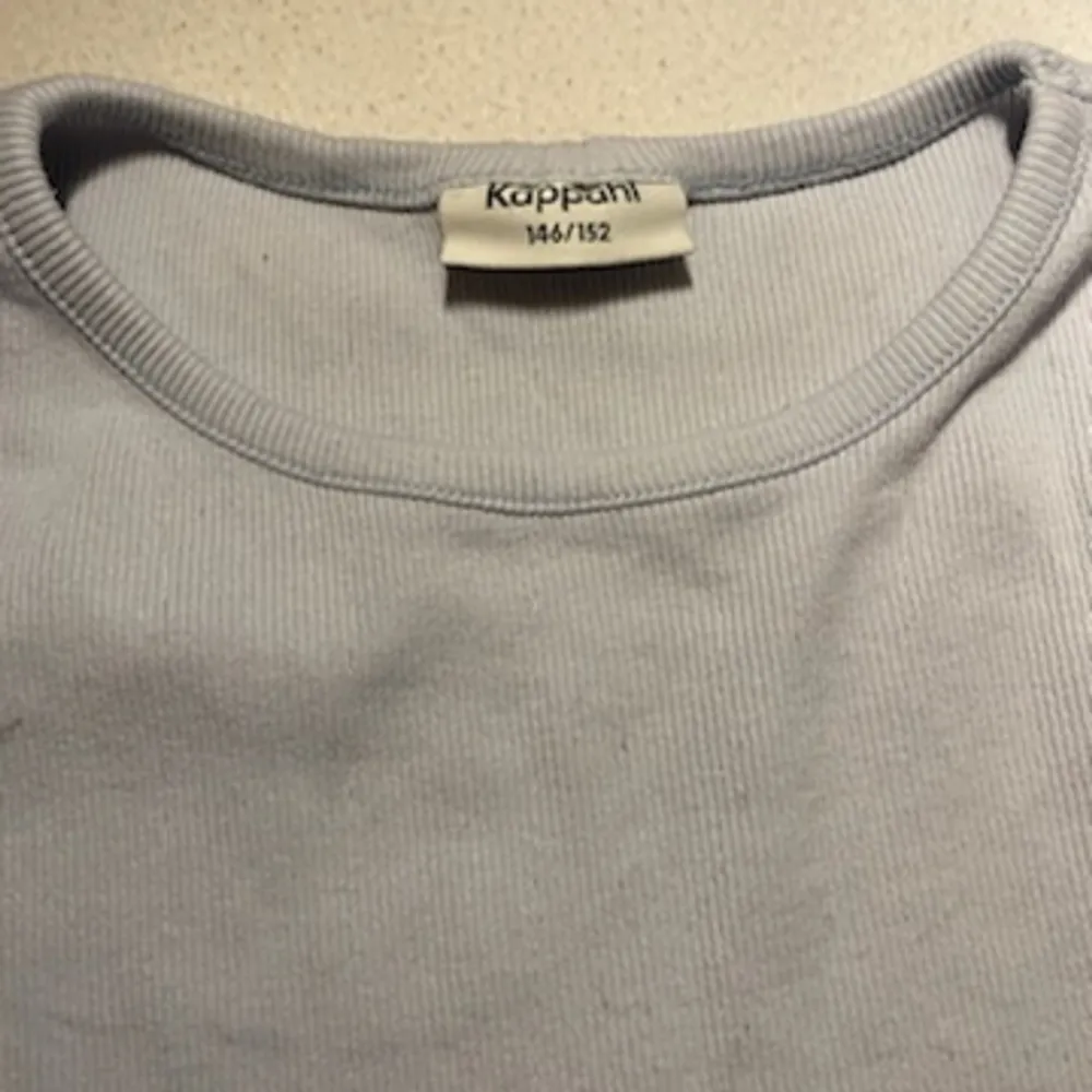 En ljusblå t-shirt med små ränder i tyget. Från KappAhl och storlek 146-152. Ganska kort i magen, men inte riktigt lika kort som en magtröja.. T-paidat.