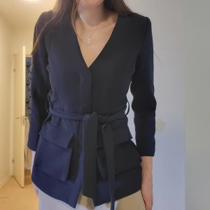 Jacka från YAS - Ny jacka från YAS i mörkblått. Bälte i midjan, dolda knappar framtill och lite smått "ribbad" textur. I nyskick och knappt använd. Svarar på alla frågor.