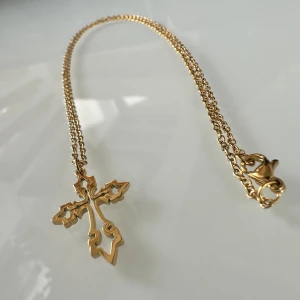 Kors halsband  - Ett klassiskt kors, här med lite rundade former, är fint att bära och ge bort som gåva. Halsbandet har en elegant, lite långsmal kedjelänk som håller korset och ses här i blank 14K guldplätering. 
