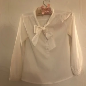 Vacker blus - Vit blus med knappar och rosett framtill. Rosetten går att knyta så som man vill ha den. Fin baksida. Är inte använd många gånger så är i fint skick, dock hittade jag någon liten fläck på insidan av ena ärmen, syns knappt då de är så små. Köpt på Åhléns. 