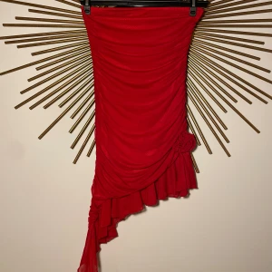 Röd axelbandslös klänning - En elegant röd axelbandslös klänning med en tight passform. Klänningen har en volangdetalj vid fållen och en dekorativ blomma på sidan. Endast använd en gång på en fest! Slutsåld på hemsidan! ❤️❤️💃