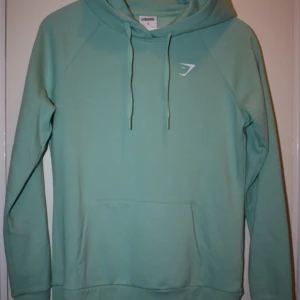 Hoodie Gymshark - Armhåla till armhåla = 50cm Armhåla till handled = 55cm Axel till handled = 64cm Axel till axel = 46cm Nacke till midja = 65 cm  Märke: Gymshark Material: Bomull och Polyester 