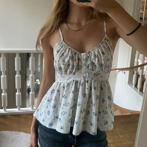 Vit blommig linnetopp - Vit linnetopp med blå blommigt mönster. Volang vid nederkanten. Har knappar framtill❣️ 