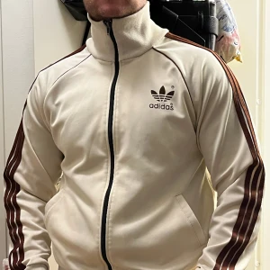 Adidas - En adidas ziptröja, storlek M. Beige i färgen. Fin tröja men en lite fläck på ena ärmen. Går ty fixa om man vill. Använder inte så säljer billigt!