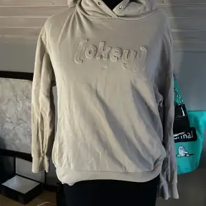 En beige hoodie från Lindex med texten 'okey' i relief på framsidan. Hoodien har långa ärmar och en ribbstickad nederkant samt muddar vid ärmslut.