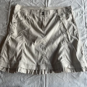 Beige kjol från Coayoun - En beige kjol  med volangdetaljer vid nederkanten. Kjolen har två framfickor och två bakfickor samt knappar och dragkedja framtill.