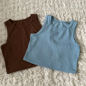 Två ribbade toppar - Två ribbade toppar i olika färger. Den ena är brun och den andra är ljusblå. Båda topparna är ärmlösa och har en croppad design. Båda 2 för 40kr. 