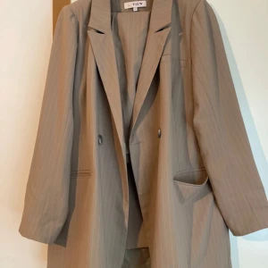 Beige kavaj från A View - En elegant beige kavaj från A View med diskreta ränder. Kavajen har en klassisk design med två knappar framtill och fickor på sidorna. Den är tillverkad i ett lätt och bekvämt material, perfekt för både formella och semi-formella tillfällen.