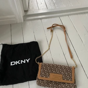 DKNY handväska  - Helt ny DKNY handväska i brunt/beige skinn och beige tyg. Justerbar axelrem så väskan kan bäras cross body eller på ena axeln. Fack med dragkedja inne samt två mindre fack förutom större.  Svart dust bag medföljer. Nypris ca 1500 kr. 
