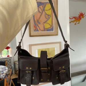 Brun axelväska i skinn - En brun axelväska i skinn med flera fack och detaljer i metall. Väskan har en justerbar axelrem och stängs med ett spänne framtill. Insidan är fodrad med svart tyg och har flera mindre fack för förvaring.💓💓Fint skick!