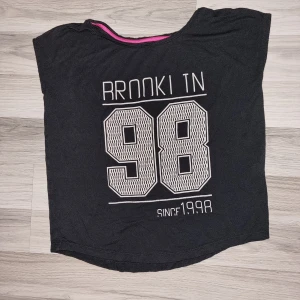 Svart t-shirt med tryck - En svart t-shirt med tryck 'RROOKI TN 98 SINCE 1998' i vitt. T-shirten har korta ärmar och en rund halsringning. Från ellos i stl 152