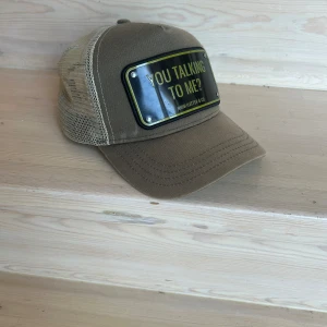 Beige keps från John Hatter & Co - En beige keps från John Hatter & Co med texten 'YOU TALKING TO ME?' på framsidan. Lätt solblekt. Kepsen har ett nätmaterial på baksidan för bättre ventilation och en böjd skärm.