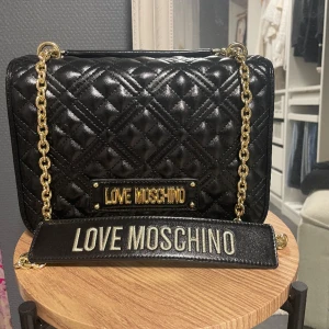 Moschino Väska  - Helt oanvänd Moschino väska! Längd: 33 cm Höjd: 23,5  Djup: 12, 5 cm . Det finns tre fack och en med dragkedja. Väskan stängs med magnet knapp 🥰🥰 Köpt för ca 3000kr 