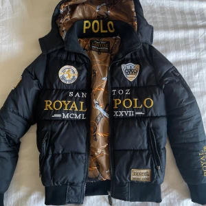 Polo santoz jacka - Varm och skön jacka i svart och guldig färg. Hela insidan med matchande guldigt mönster, och yttersidan med flera broderier och patches i samma matchande guldiga färg. Avtagbar luva med dragkedja. Två sidfickor med dragkedja, samt en innerficka