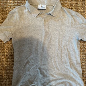 Grå pikétröja från The Shirt Factory - En grå pikétröja från The Shirt Factory. Tröjan har korta ärmar och en klassisk krage med knappar.