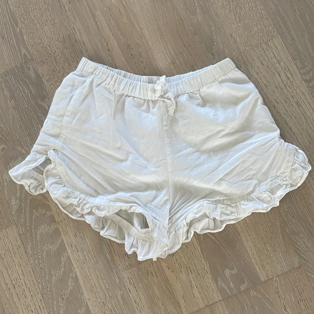 Ett par vita shorts med elastisk midja och volangdetaljer vid bensluten. Shortsen har en knytning framtill som prydnad runt midjan. Storlek står inte men åldern är 8-9 år, man kan ha det som vanliga short eller som pjamas/mys shorts 😀. Shorts.