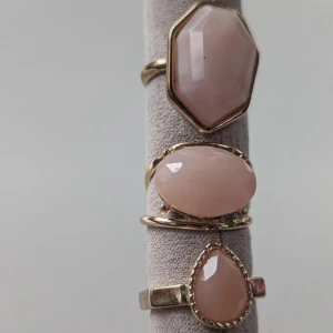 Set med tre ringar med rosa stenar - Ett set med tre eleganta ringar, var och en prydd med en rosa sten. Den första ringen har en stor, åttakantig sten, den andra en oval sten och den tredje en droppformad sten. Alla ringar har en guldfärgad bas.