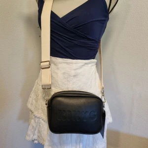 Svart axelväska från Kirys - En stilren svart axelväska från Kikys med präglad logotyp på framsidan. Väskan har en justerbar axelrem i beige färg och dragkedja för säker förslutning. Perfekt för vardagligt bruk.