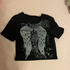 Svart croppad topp med tryck - En svart croppad topp med korta ärmar och ett detaljerat tryck av vingar och blommor i grått och vitt på framsidan. Toppen har en rund halsringning och är tillverkad i ett mjukt material. Skriv gärna om ni har frågor 💕💕
