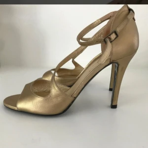 Högklackade skor Guld Äkta läder Rizzo Stl 38 - Snygga guld skor med hög klack. Stl 38. Rizzo. Äkta läder. Använda en gång, inga defekter, syns lite nött på sulan och lite svarta prickar på innersulan. Klackhöjd mitt bak 10 cm. Tillverkade i Spanien. Nypris 900kr