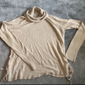 Beige polotröja med snörning - En beige polotröja från Love Knitwear med långa ärmar och ribbstickade muddar vid ärmslut och nederkant. Tröjan har en polokrage och snörningsdetaljer på sidorna.