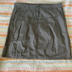 Grå kjol från Soyaconcept - En grågrön kjol från Soyaconcept med broderade detaljer på vänster sida. Kjolen har två framfickor och två bakfickor med knappar. Den har en knapp och dragkedja framtill.