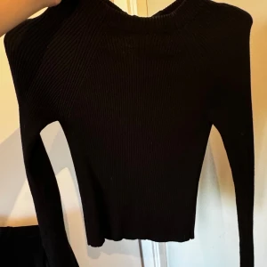 Svart ribbad tröja med öppen rygg - En svart ribbad tröja med långärmad design och öppen rygg. Tröjan har en knytning i nacken och är tillverkad i ett stretchigt material.