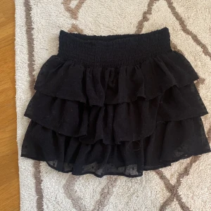 Svart volangkjol från Lindex - En svart kortkjol från Lindex med volangdetaljer. Kjolen har en elastisk midja och är tillverkad i ett lätt och luftigt material. den är i storlek 134/140 men är väldigt stretchig så passar mig som är 170cm, men den blir lite kortare☺️