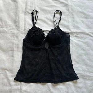 Svart korsett med spetsdetaljer - En elegant svart korsett med spetsdetaljer och justerbara axelband. Korsetten har en genomskinlig mesh-design och dekorativa blommor på kuporna.
