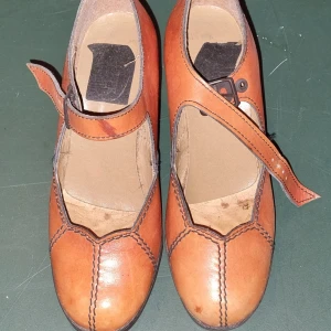 Bruna skor med klack och rem - Ett par bruna skor med klack och rem över foten. Skorna har en rund tå och är tillverkade i skinn. De har en dekorativ söm längs kanterna och en justerbar rem med spänne. Skinn