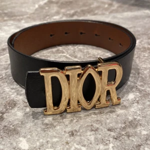 Svart skinnbälte från Dior - Ett elegant svart skinnbälte från Dior med ett stort guldfärgat spänne som stavar 'DIOR'. Bältet har flera hål för justerbar passform.