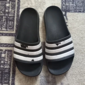 Adidas tofflor - Säljer mina Adidas tofflor. De är lite slitna som man kan se och på baksidan av skorna finns det en inbuckla typ, se bild 3. Men det påverkar inte bekvämligheten alls. Det står ingen storlek på, men skulle gissa på 38 1/2. Köparen står för frakten! 