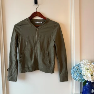 Olivgrön kofta från 157 - Olivgrön kofta med bomberjacka stil, med rund hals och dragkedja framtill. Jackan har två sidofickor och är tillverkad i mjuk bomull. Enkel och clean design som funkar till många olika stilar. Långärmad och med normal passform.