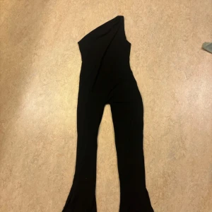 Svart byxdress från Oh Polly. PETITE - Säljer en svart, snygg byxdress från Oh Polly med asymmetrisk axel och utsvängda ben. Materialet är mjukt och stretchigt, vilket gör den riktigt bekväm. Perfekt för dig som gillar en trendig och cool look med lite extra edge.