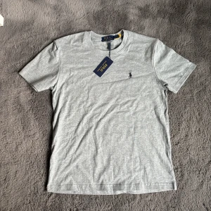 Grå t-shirt från Polo Ralph Lauren - Snygg grå t-shirt från Polo Ralph Lauren med klassisk rund hals och broderad svart logga på bröstet. Tillverkad i mjuk bomull för en skön känsla. Perfekt basic-plagg med stilren look och normal passform.