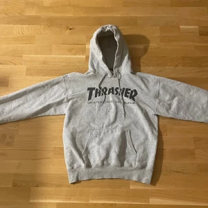 Grå Thrasher hoodie med tryck - Säljer en grå hoodie från Thrasher med klassiskt svart logotryck på bröstet. Tröjan har huva med snörning och en stor magficka. Perfekt för dig som gillar streetwear och skatekultur. Mjuk och bekväm bomullsmix som passar till chill dagar.