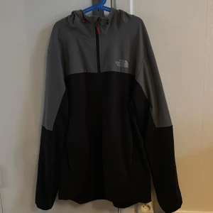 Svart och grå vindjacka från The North Face - Snygg vindjacka från The North Face i svart och mörkgrått med huva och justerbara snören. Jackan har en half zip-dragkedja framtill och reflekterande logga på bröstet. Perfekt för dig som gillar stilren och funktionell design.