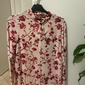 Blommig långärmad blus från Stockh LM - Säljer en snygg blus från Stockh LM med blommigt rött mönster på ljusbeige botten. Blusen har lång ärm och en liten knytning vid halsen som ger en extra detalj. Perfekt för dig som gillar stilrena och trendiga plagg med lite extra attityd.
