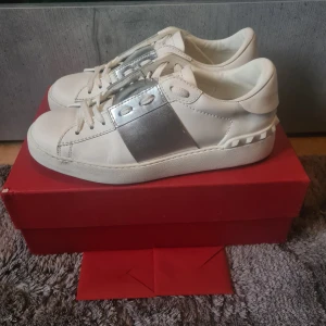 Valentino Garavani - Snygga Valentino Garavani Open sneakers i vitt skinn med metallic-silver rem. De är I storlek 37,5 och är I bra skick. De har blivit fixade lite på det silvriga. Hör av dig vid frågor.