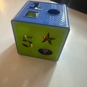 klosslåda - Bilden visar en färgglad formsorterare i plast, avsedd för små barn. Produkten har olika former och siffror på sidorna där barn kan stoppa in matchande klossar. Inga synliga defekter eller slitage, ser ut att vara i gott skick. Perfekt för att utveckla motorik och igenkänning av former.