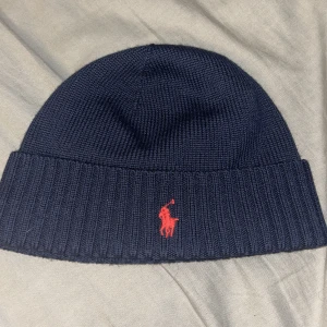 Mörkblå stickad mössa från Polo Ralph Lauren - Snygg mörkblå stickad mössa från Polo Ralph Lauren med klassisk röd broderad logga framtill. Mössan har bred ribbad kant och är perfekt för kyliga dagar. Enkel och stilren design som passar till det mesta.