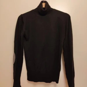 Svart polotröja med gråa armbågslappar - Svart polotröja med lång ärm och gråa armbågslappar. Tröjan har en klassisk polokrage och är tillverkad i ett mjukt stickat material som känns skönt mot huden. Perfekt för dig som gillar stilrena och enkla plagg med en liten twist.