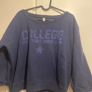 Blå college-tröja med tryck, stl L - Mörkblå sweatshirt från College Outfitters med stort tryck 'COLLEGE OUTFITTERS SINCE 1979' och en stjärna på bröstet. Tröjan har bred halsringning och är gjord i mjuk bomull. Lite kort modell i midjan. Perfekt för chill dagar och enkel att matcha med jeans.