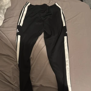 Svarta Adidas mjukisbyxor st M - Svarta mjukisbyxor från Adidas med klassiska vita ränder längs sidorna och Adidas-logga på båda benen. Byxorna har elastisk midja och är gjorda i mjukt syntetmaterial, perfekta för chill eller träning.