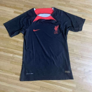 Liverpool FC svart Nike fotbollströja - Snygg svart fotbollströja från Nike med Liverpool FC-logga och röd Nike-swoosh på bröstet. Tröjan har korta ärmar, röd detalj vid halsen och är tillverkad i ett lätt, ventilerande material med Dri-FIT-teknologi. Perfekt för dig som vill representera LFC på planen.